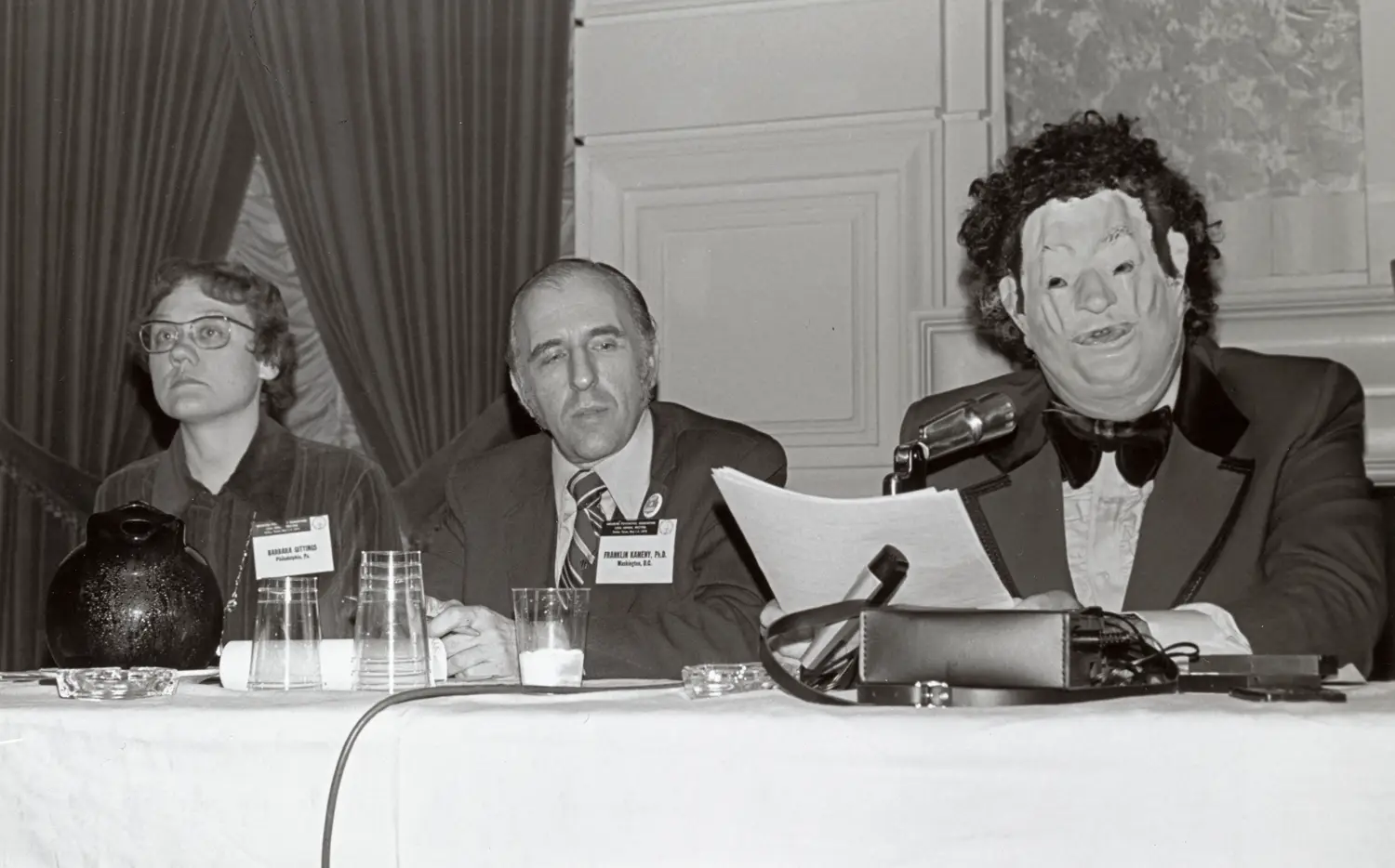 From left to right, a woman, man and person wearing a wig and mask are seated at a conference table.