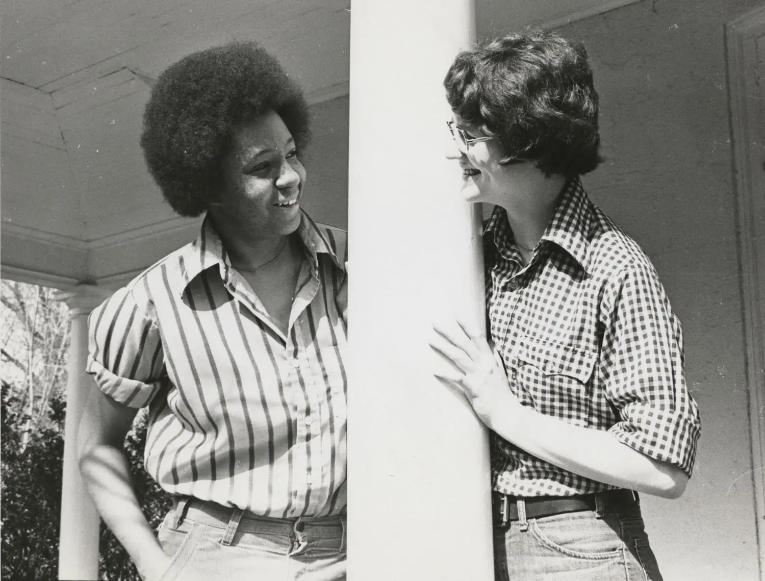 Two women are leaning on a pillar, smiling at each other.